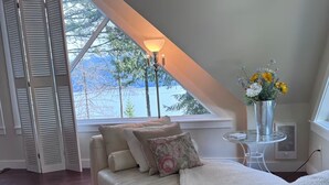 Une pièce avec un plafond en pente, un canapé avec des coussins, une table en verre, un vase rempli de fleurs et une vue sur les arbres et l’eau à l’extérieur.