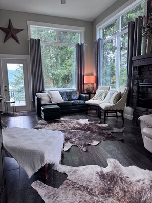 A spacious living room with a large window, a fireplace, a leather sofa, and a unique cowhide rug. - Luxe two-storey home provides a beautiful modern unique romantic getaway! (Anglemont)