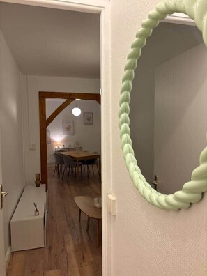 Une pièce avec une porte en bois, un miroir vert pâle à motifs tourbillonnants, un radiateur blanc et un coin repas comprenant une table et des chaises en bois.