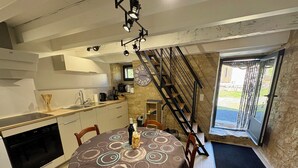 A dining area with a round table, chairs, a kitchen with a stove, and a staircase leading to an outdoor area. - The Pechplazen grangette (Rudelle)