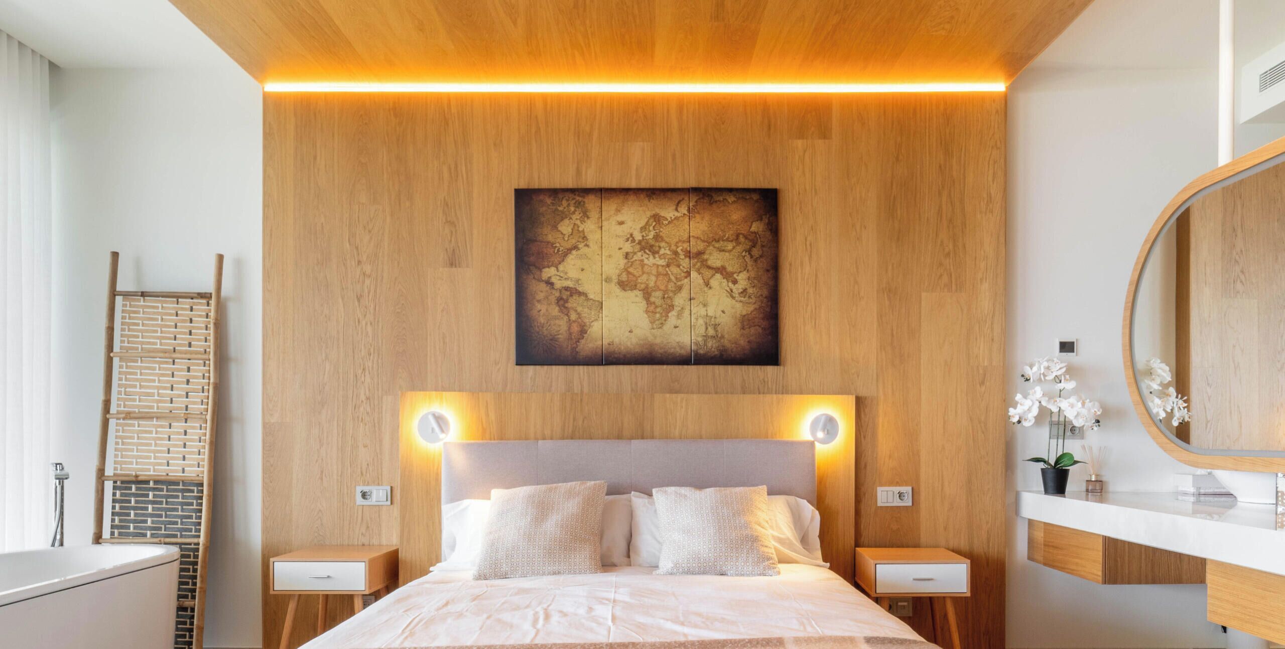 Une chambre d’hôtel moderne avec une tête de lit en bois, une œuvre d’art représentant une carte du monde, un miroir rond et une table de chevet avec une lampe.