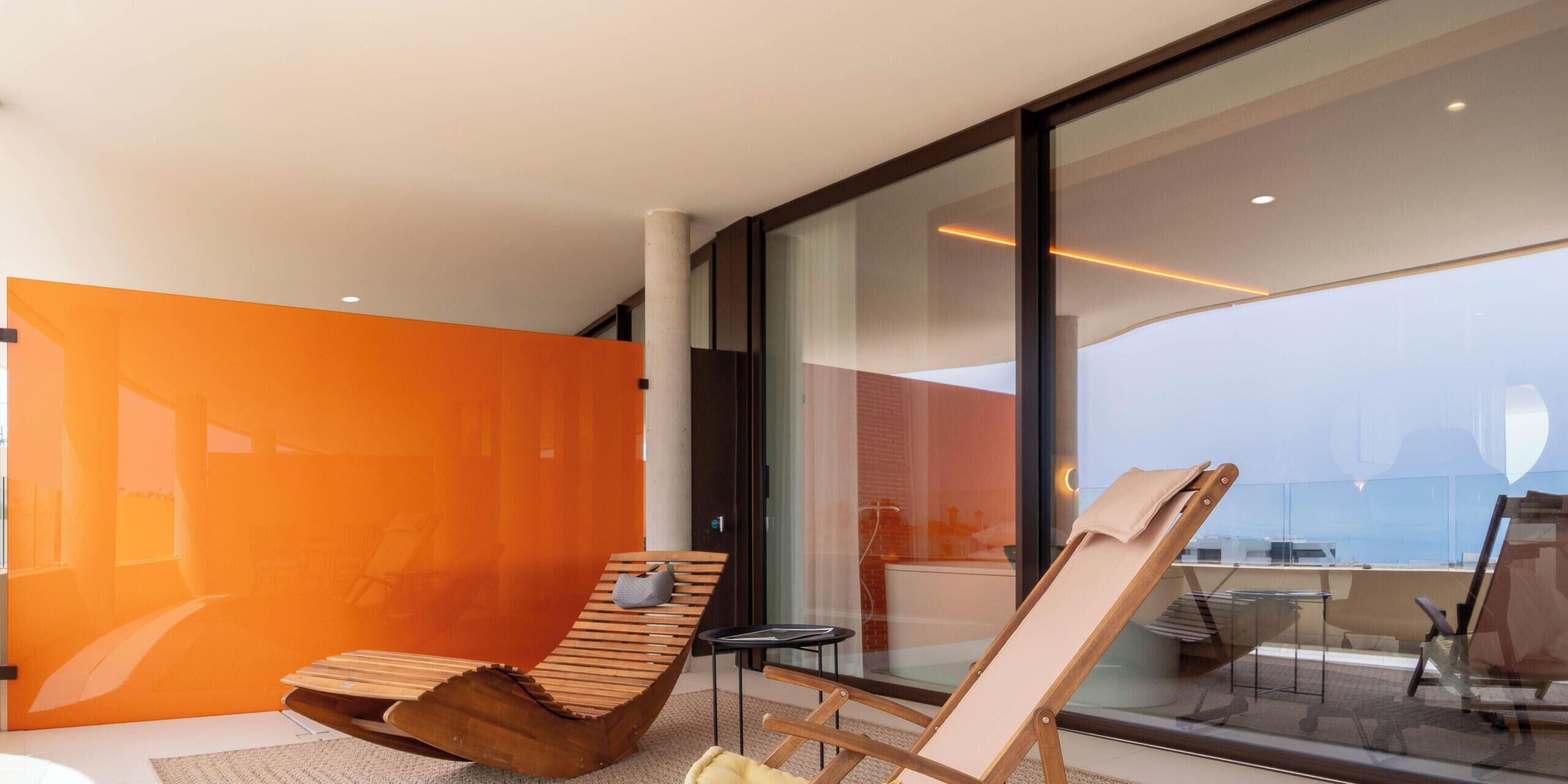 Una habitación moderna con un amplio ventanal, una silla de madera, una mesita pequeña y una pared de acento color naranja brillante.