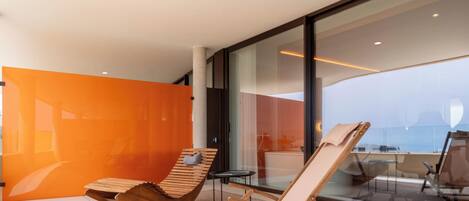 Una habitación moderna con un amplio ventanal, una silla de madera, una mesita pequeña y una pared de acento color naranja brillante.