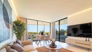 Un salón moderno con un televisor de pantalla plana grande, un sofá, una zona de comedor con sillas y un ventanal que ofrece vistas al verdor.