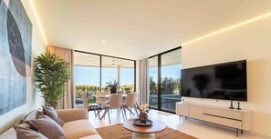 Ein modernes Wohnzimmer mit einem großen Flachbildfernseher, einer Couch, einem Essbereich mit Stühlen und einem großen Fenster mit Blick auf Grünflächen.
