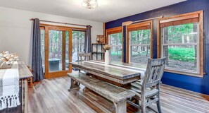 A dining area with a wooden table and benches, large windows with forest views, and a ceiling light fixture. - Pine Loch, cozy cabin sleeps 9 with 3 bedrooms, 2 baths. Gas fire pit on deck. (Pocono Lake)