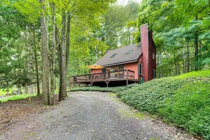 A red cabin with a balcony, surrounded by trees and a gravel path. - Pine Loch, cozy cabin sleeps 9 with 3 bedrooms, 2 baths. Gas fire pit on deck. (Pocono Lake)