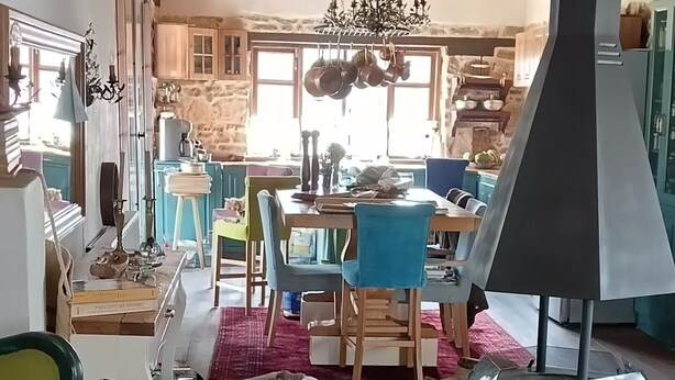 A rustic interior with a wooden ceiling, a dining area with a table and chairs, a fireplace, and a large window.