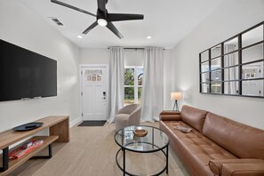 A modern living room with a flat-screen TV, a leather sofa, a glass coffee table, and a ceiling fan.