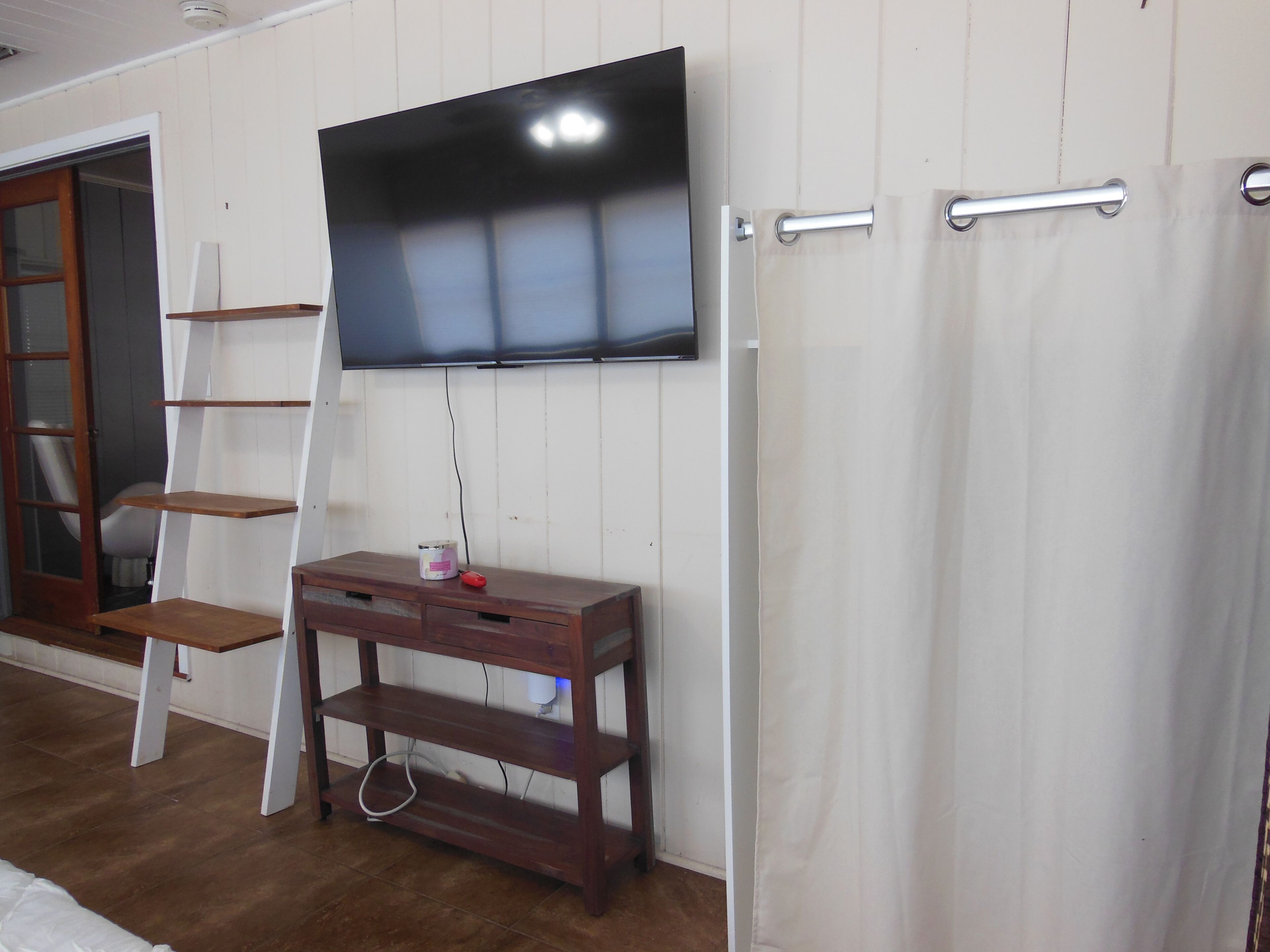 Quarto com televisão na parede, mesa de console de madeira, cortina branca e escada de madeira.