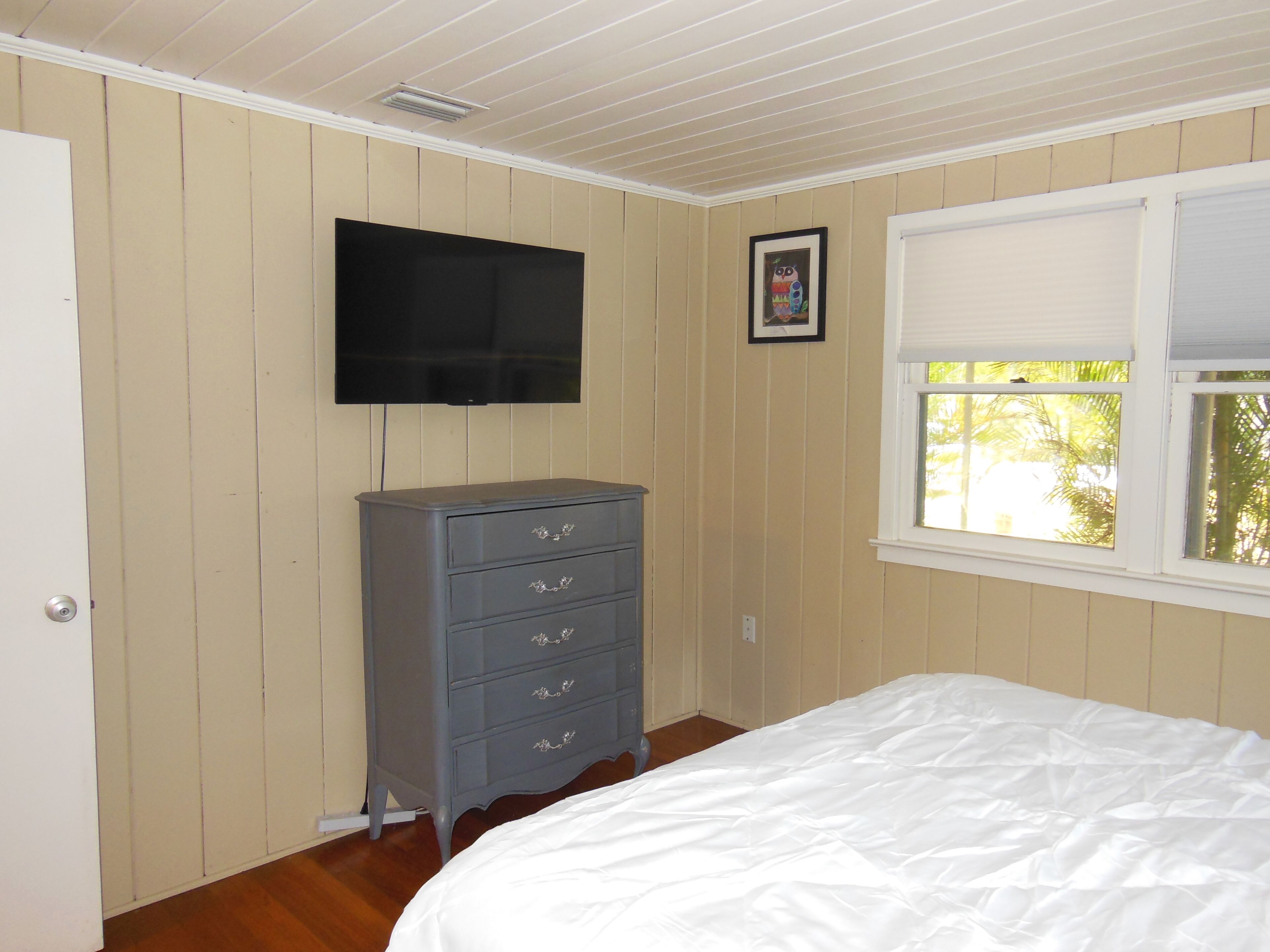 Een slaapkamer met een bed, een dressoir, een aan de muur bevestigde televisie en een raam met zonwering.