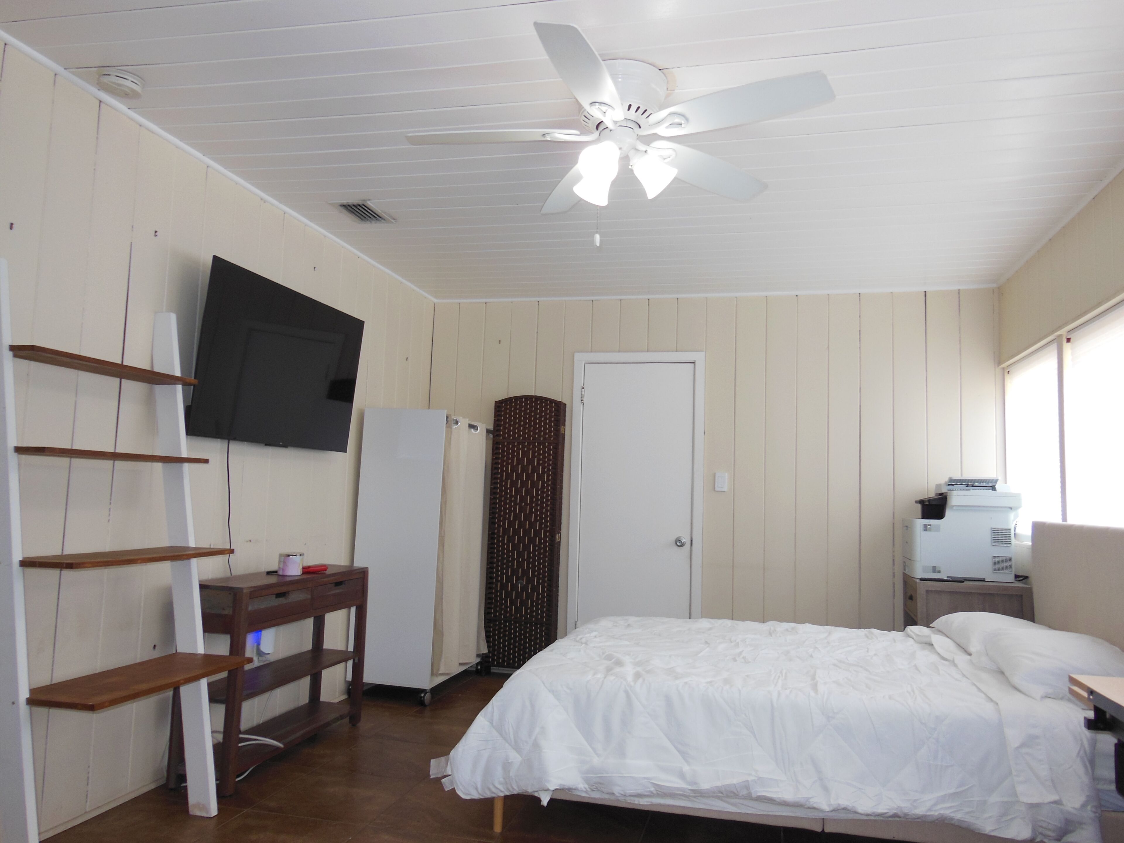 Un dormitorio con cama, escritorio, televisor, puerta y ventilador de techo.