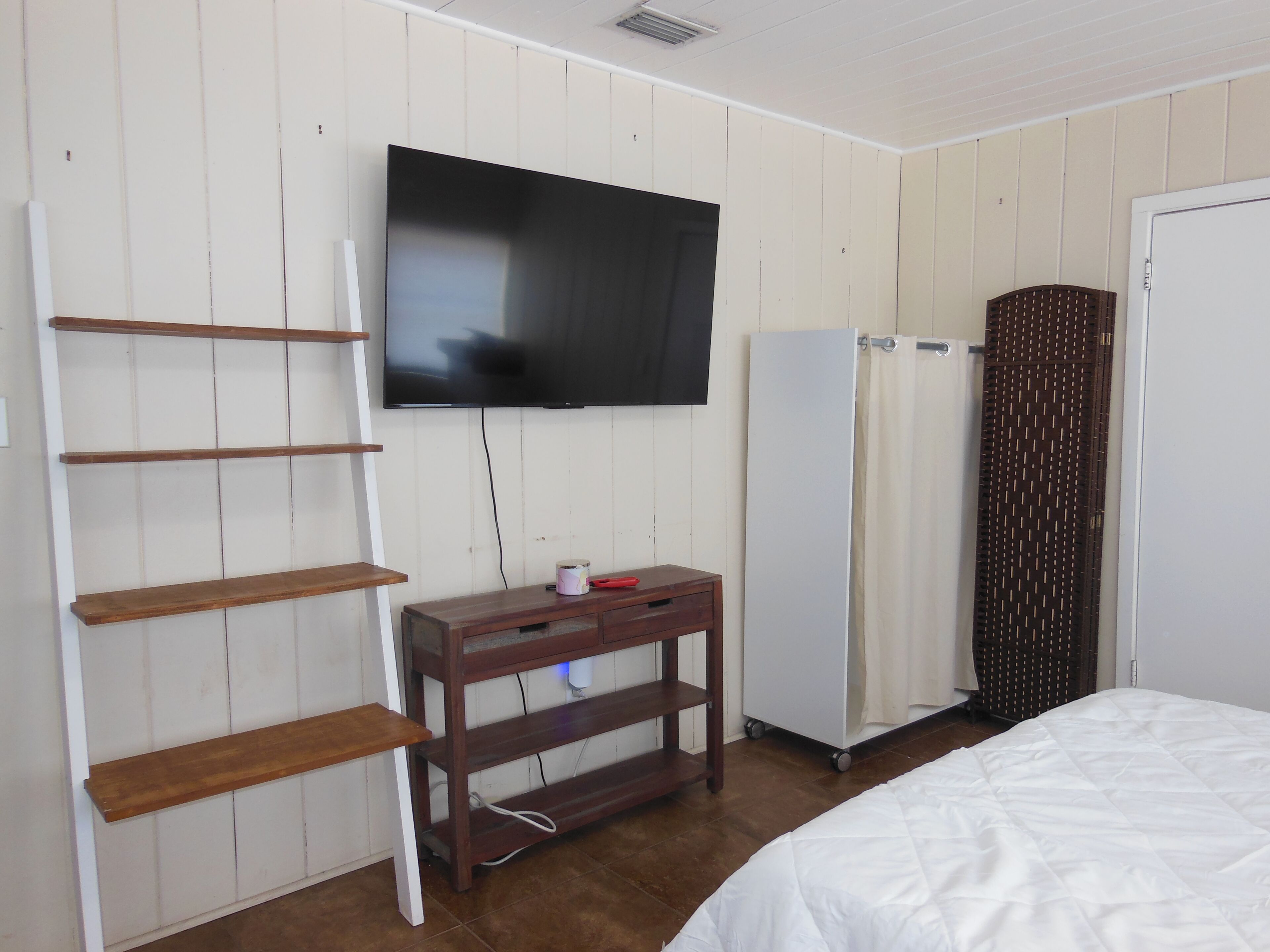 Una habitación con una televisión montada en la pared, estantes de madera, una mesita de madera pequeña, una cama blanca y un armario blanco.