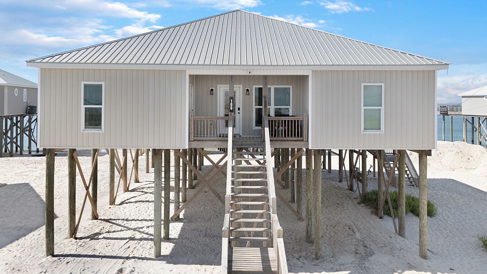 Une maison de plage avec un escalier central menant à une terrasse, le tout entouré de sable.
