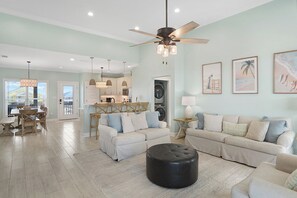 A spacious living room with a large sofa, a coffee table, and a dining area with a table and chairs. There is a kitchen in the background with cabinets and a ceiling fan. - Beachfront + Private Hot Tub, Pet Friendly, Hammock + Swings, Loaded, Sleeps 12 (Dauphin Island)