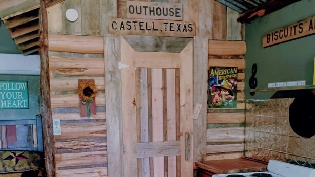 Eine rustikale Küche mit einer Holztür, auf der 'Outhouse Castell, Texas' steht, einem weißen Herd und einem roten Schrank.