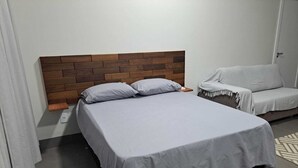 A bedroom with a bed, a wooden headboard, a nightstand, a grey sofa, and a patterned rug. - LU'S HOUSE APARTMENTS (102) (Primavera do Leste)