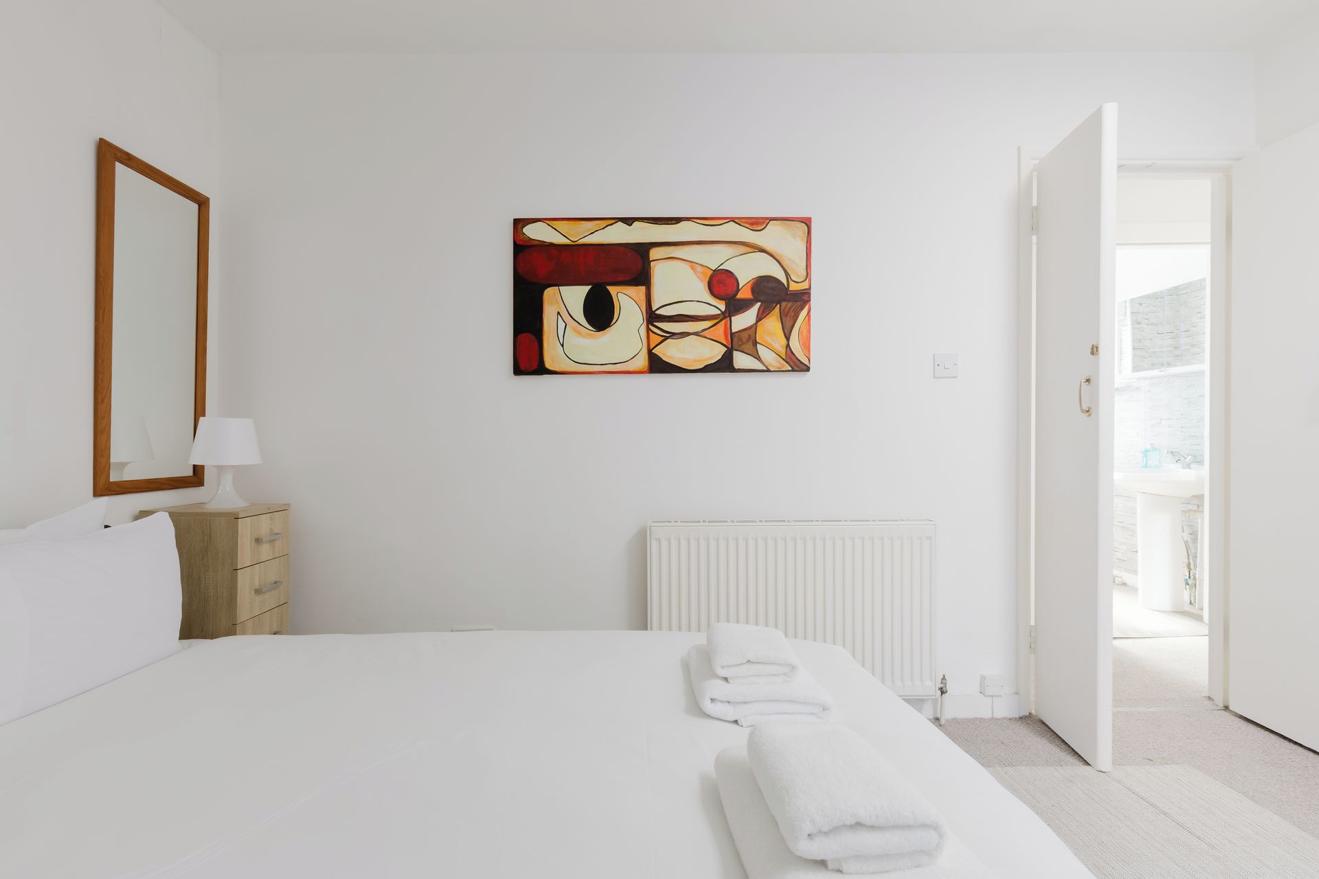 Une chambre d’hôtel avec un lit, un radiateur, un tableau, une commode en bois et une porte donnant sur un balcon.