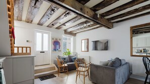A modern living room with a grey sofa, wooden chairs, a TV mounted on the wall, and a kitchen area with a radiator. - The Beatles Inspired 3BDR Home by Abbey Road (Greater London)