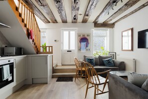 Une cuisine moderne équipée d’un four à micro-ondes, un coin repas avec des chaises en bois, un canapé gris et un escalier avec des poutres apparentes.