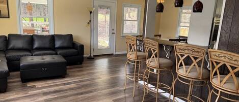 A living room with a sectional sofa, a dining area with bar stools, and a kitchen with pendant lights.
