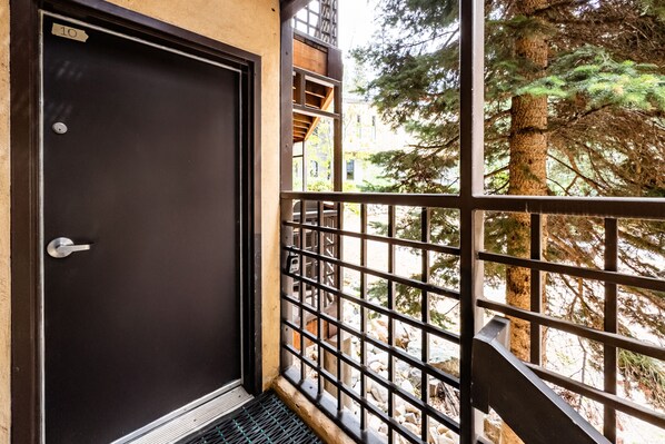 A door with a metal handle and a number sign, a metal railing, and a view of trees and a building in the background.