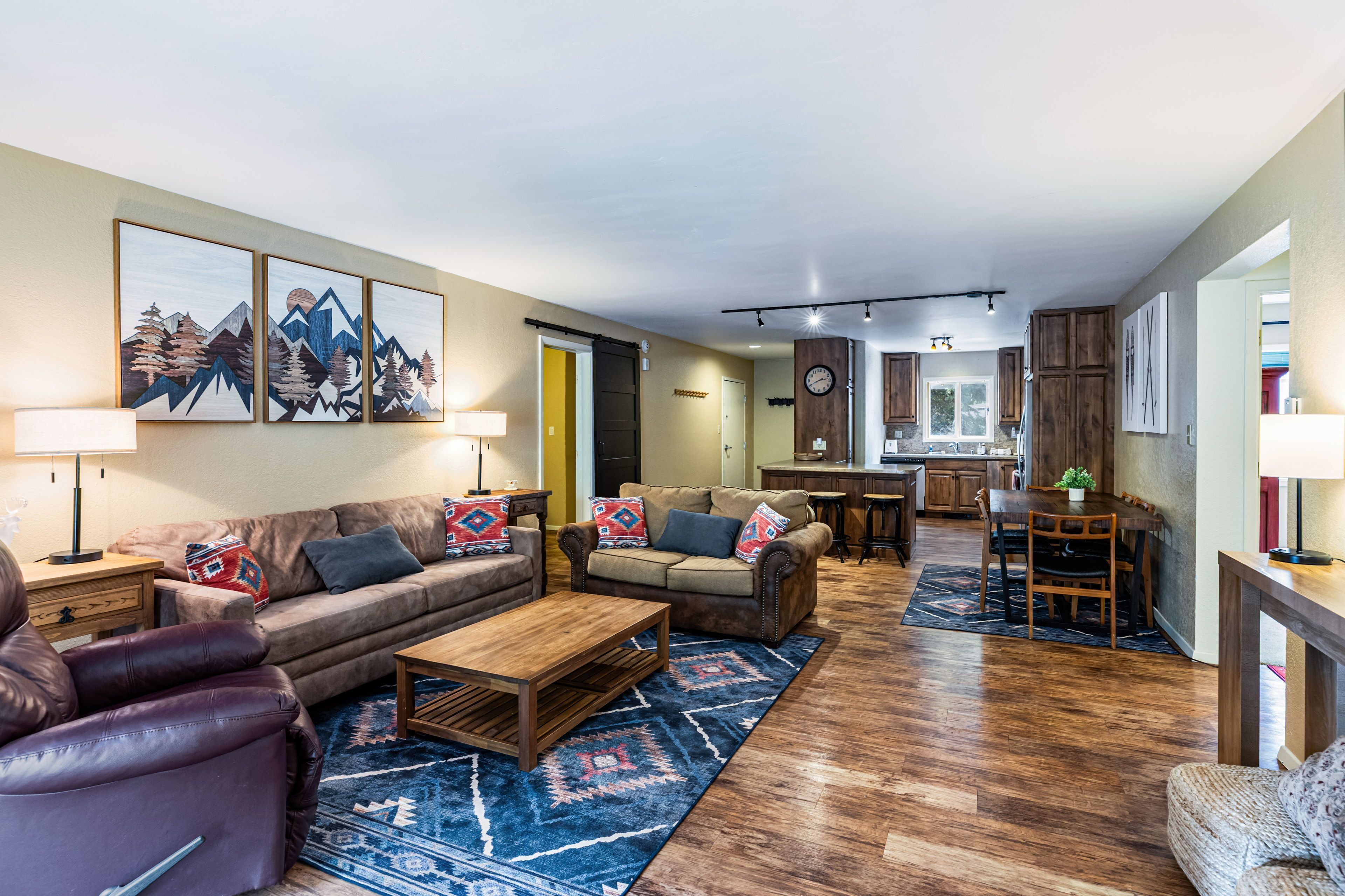 Amplia sala de estar con sofá, sillones y una mesa de centro. Se ve una cocina con muebles de madera y un comedor con mesa y sillas al fondo.