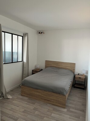 A bedroom with a bed, a nightstand, a window with curtains, and a wall-mounted light fixture. - Charming T3 - Garage - Balcony (Meyzieu)