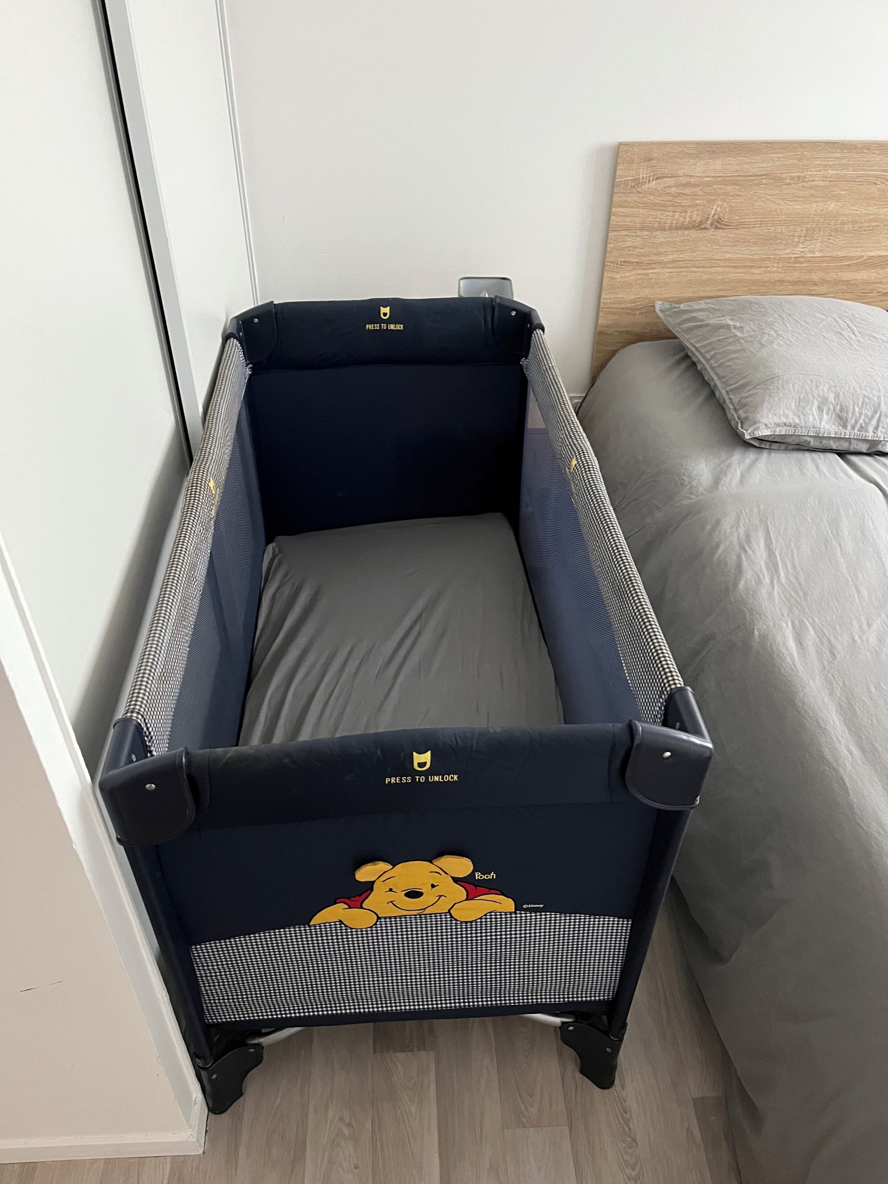 Een compacte, donkerkleurige koffer met een cartoon ontwerp en een 'Press to Unlock' label, geplaatst naast een bed met een grijze kussen en een houten hoofdbord.