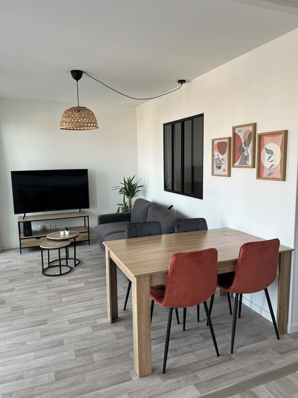 Ein modernes Wohnzimmer mit Flachbildfernseher, einem grauen Sofa, einem Holzesstisch mit roten Stühlen und einem großen Fenster.