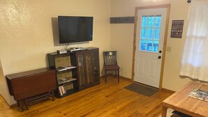 A living room with a wooden floor, a television mounted on the wall, a cabinet with glass doors, a door with a window, and a small table with magazines.
