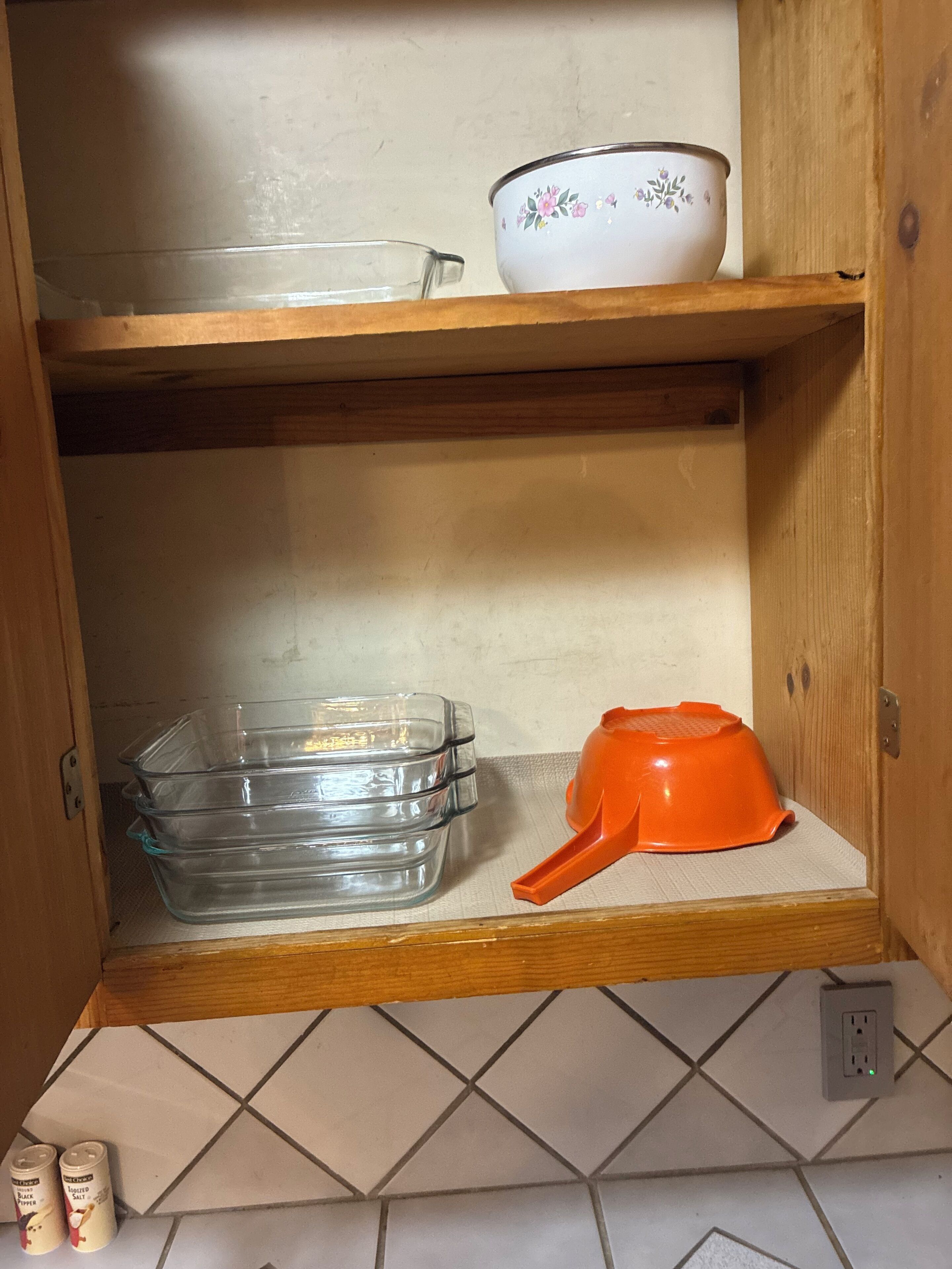 A kitchen cabinet with a clear glass dish rack, a white bowl with floral patterns, and an orange measuring cup.