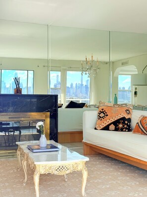 Ein Wohnzimmer mit Klavier, einer gepolsterten Couch mit gemusterten Kissen, einer Kristallleuchtre und Blick auf eine Stadtlandschaft durch große Fenster.