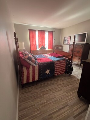 Une chambre à coucher avec un lit, une commode, un téléviseur et une fenêtre avec des rideaux.