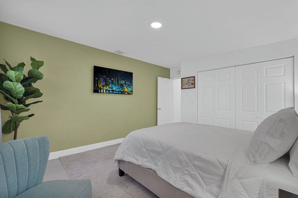 A bedroom with a bed, a chair, a plant, and a cityscape painting on the wall.