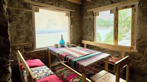 A rustic stone room with a bed, a table, and two windows.