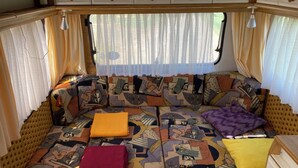 Un intérieur de camping-car chaleureux, avec un canapé à motifs, une tête de lit en bois et une fenêtre agrémentée de rideaux légers.