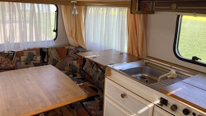 Un intérieur de camping-car compact comprenant un coin cuisine, un espace repas et une fenêtre avec des rideaux.