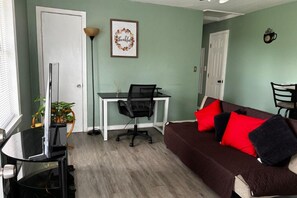 Una sala de estar con un televisor negro sobre un mueble bajo, un escritorio blanco con una silla, un sofá marrón con cojines rojos y negros, y un letrero en el pared que dice 'thankful'.