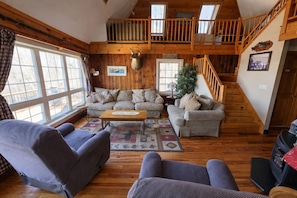 Interior acogedor de una cabaña con escalera de madera, un conjunto de sofás, una mesa de centro y una cabeza de venado decorativa.