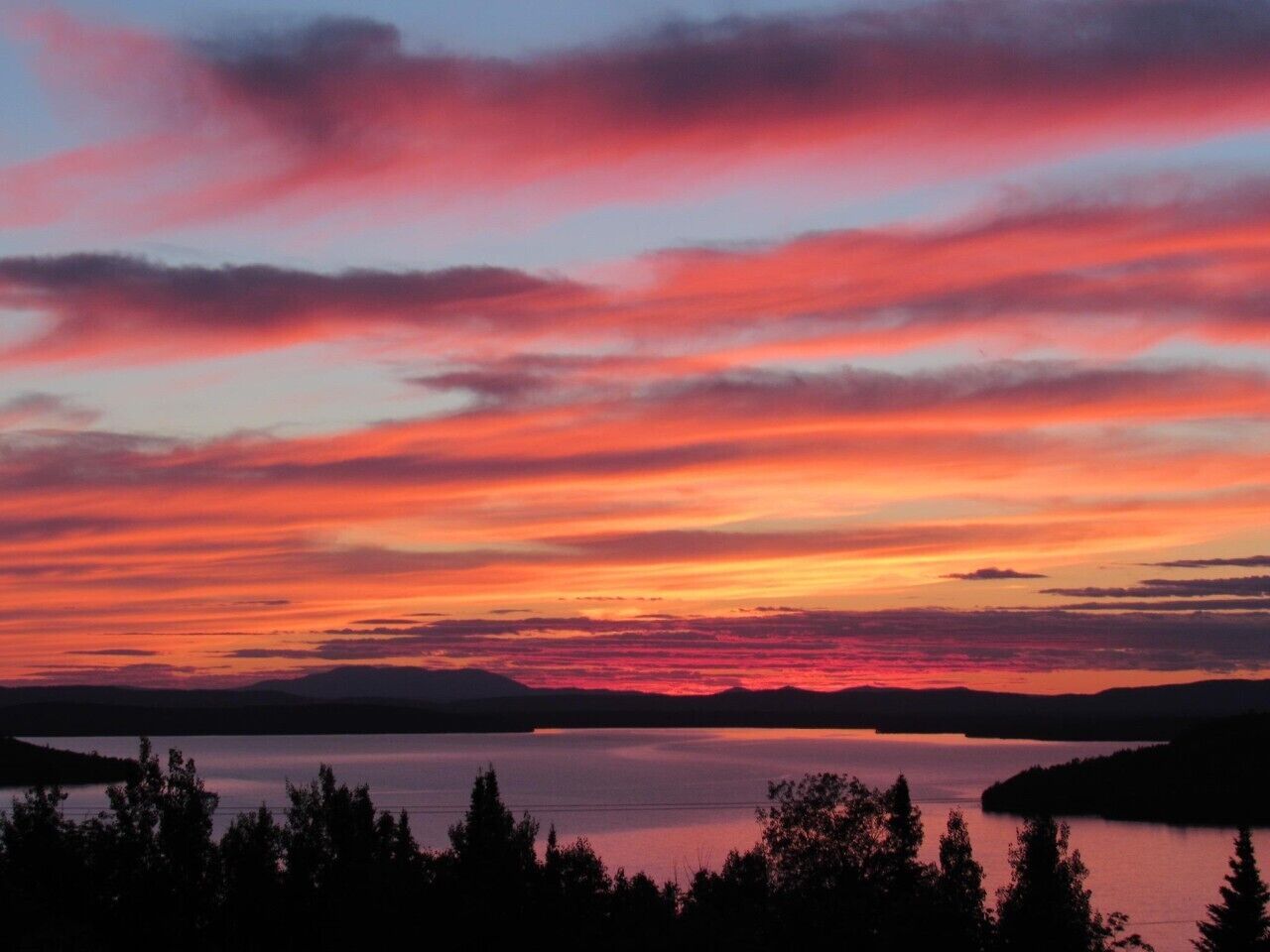 A sunset over a lake with silhouettes of trees and distant mountains.