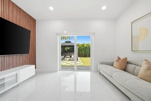 A modern living room with a flat-screen TV, a sofa, and a sliding glass door leading to an outdoor patio. - 4BR Modern Home | Sleeps 14 | BBQ & Outdoor Space (Hialeah)