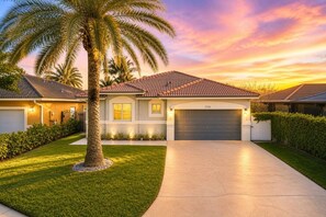 A two-story house with a tiled roof, a garage, and a well-maintained lawn with a palm tree. - 4BR Modern Home | Sleeps 14 | BBQ & Outdoor Space (Hialeah)