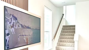 A living room with a flat-screen TV mounted on a wall, a wooden entertainment unit, a staircase, and a view of a body of water. - Brand-New- Rooftop Luxe 4BR/5BA, 15 Min to DC+ MGM (Upper Marlboro)