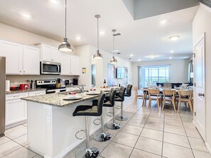 A modern kitchen with white cabinets, a granite countertop, and stainless steel appliances. There is a dining area with a table and chairs, and a large window providing a view of the outdoors. - Dream Home 7BR – Pool, Hot Tub, Game Room & Theater - Near Disney! (Davenport)