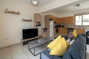 A modern living room with a flat-screen TV, sofa, coffee table, and kitchen area with wooden cabinets and a refrigerator. - 3BR Azure Pearl Seaside Villa, 50 meters from Pernera Beach in Protaras (Paralimni)