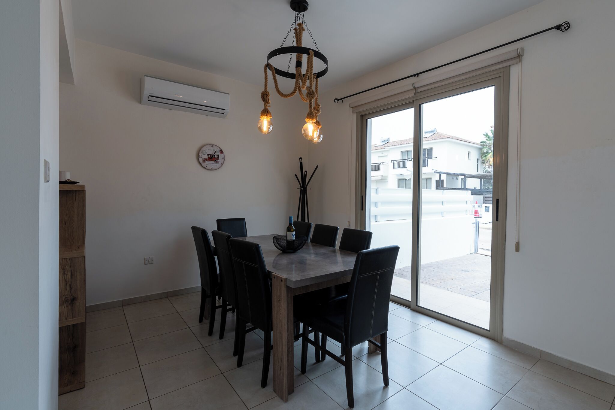 Un espace repas comprenant une table, des chaises, un lustre, un climatiseur, une horloge et une vue sur l’extérieur, accessible par une porte coulissante.