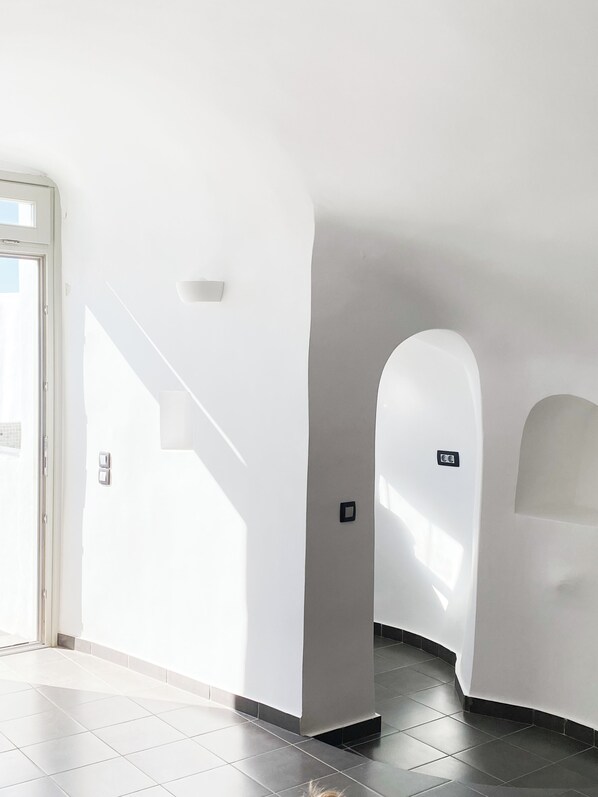 A bright hallway with white walls, a tiled floor, and a door leading outside. - Esmi Suites Santorini (Imerovigli)
