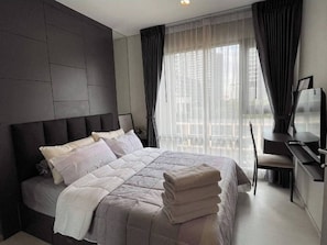 Une chambre à coucher moderne, dotée d’un grand lit, d’un bureau et offrant une vue sur la ville à travers de fines rideaux.