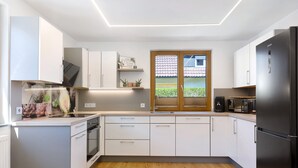 A modern kitchen with white cabinetry, a built-in oven, a refrigerator, and a window with a view of a house. - Apartment “Am Brändbach” with Private Terrace, Garden, and Wi-Fi (Bräunlingen)